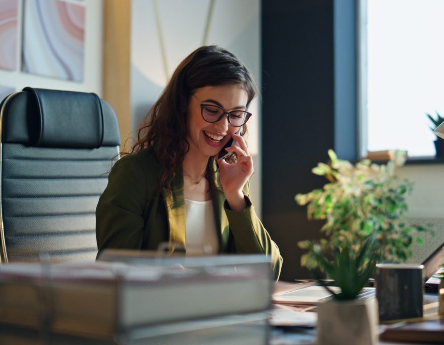 Cheerful beautiful girl office manager talking smartphone sitting modern workplace close up. Smiling business woman calling to customer offering creative company services. Girl worker looking papers.